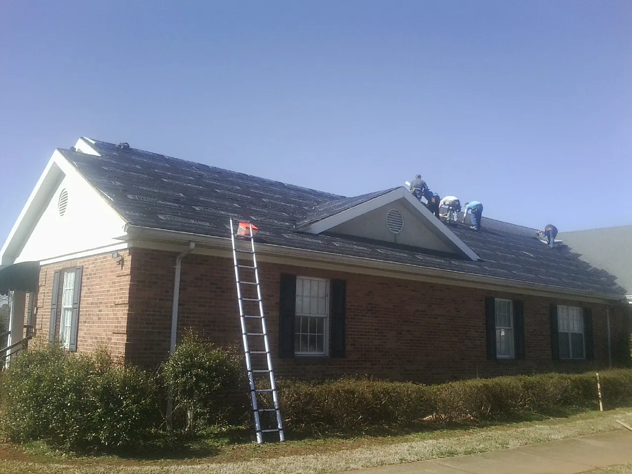 Crew tearing shingles off a brick ranch roof with a ladder set — Metal Roof Repair work by our crew in Igo