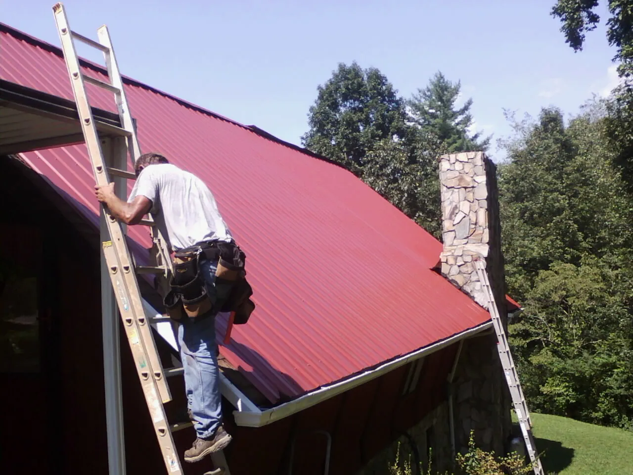 Roofer on a ladder beside a red metal roof and stone chimney — Soffit & Fascia Repair by Roofing Brothers in Igo